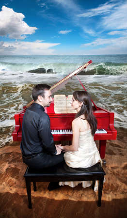 Young couple sitting in front of the red grand piano and looking at each other. Piano standing on the beach near the ocean with waves and blue sky at the background の写真素材