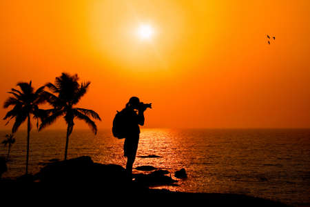 Photographer silhouette shooting sea outdoors on the rock cliff near palm trees at sunset backgroundの写真素材