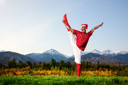 Christmas yoga utthita Hasta Padangustasana pose by happy Indian man in white trousers, red socks and Christmas hat at mountain background. Free space for textの写真素材