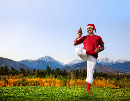 Christmas yoga vrikshasana tree pose by happy Indian man in white trousers, red socks and Christmas hat with Christmas tree at mountain background. Free space for textの写真素材