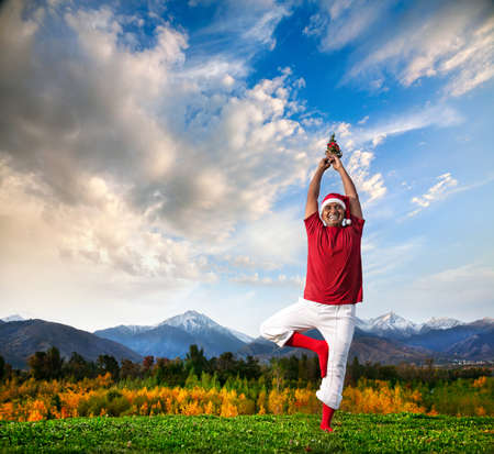 Christmas yoga vrikshasana tree pose by happy Indian man in white trousers, red socks and Christmas hat with Christmas tree at mountain background. Free space for textの写真素材