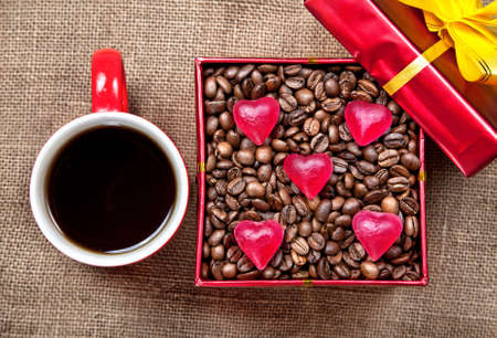 Coffee grains with heart shape candies in Red box with yellow ribbon and mug with black coffee at textured brown background on Valentines dayの写真素材