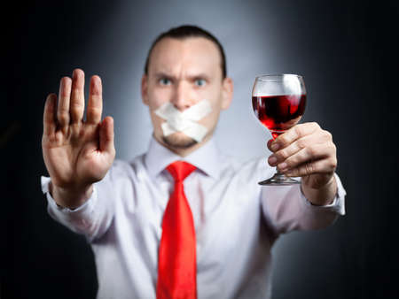 Businessman with plaster on his mouth in red tie holding the glass of red wine and gesturing stop sign by his palm at black background. Represents outcry alcoholic dependencyの写真素材