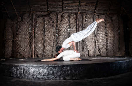 Couple yoga of woman doing vajrasana forward bending and man doing mayurasana on her back in white cloth in yoga hall, Varkala, Kerala, Indiaの写真素材