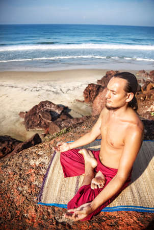 Yoga meditation in lotus pose by man with long hair in red trousers on the cliff near the ocean in Varkala, Kerala, Indiaの写真素材