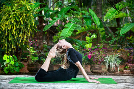 Yoga Raja Kapotasana backward bending pose by woman in black cloth in the garden with palms, banana...