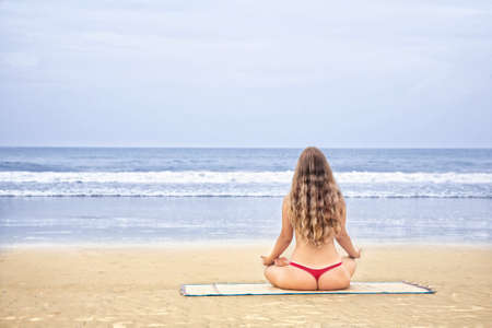 Young woman in red bikini doing meditation on the beach at ocean background. Free space for your text の写真素材