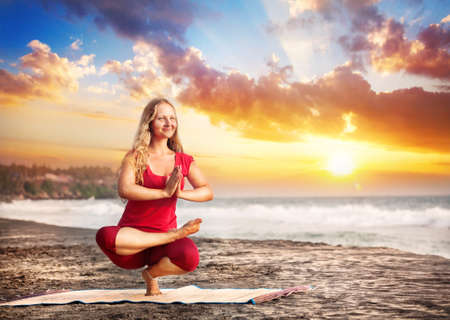 Yoga balancing pose by young woman with long hair in red cloth on the beach near the ocean at dramatic sunset background の写真素材