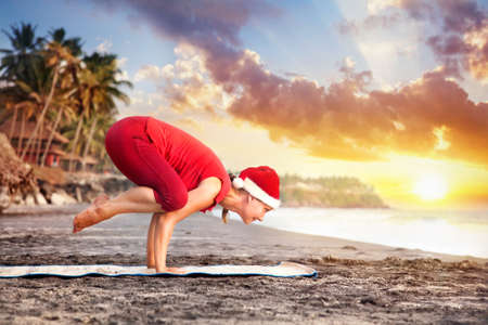 Yoga bakasana crane pose by young woman in red costume and red christmas hat on the beach near the ocean at sunset background in India の写真素材