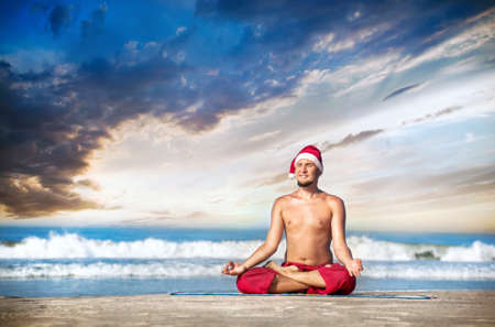 Christmas yoga meditation in padmasana lotus pose by man in red trousers and Christmas hat on the beach near the ocean in India の写真素材