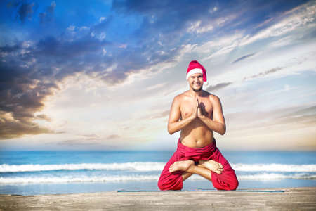 Christmas yoga in padmasana lotus pose by happy man in red trousers and Christmas hat on the beach near the ocean in India の写真素材