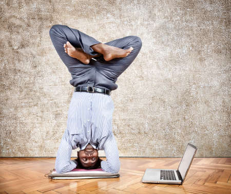 Indian businessman doing yoga headstand pose and looking at his laptop in the office at brown textured backgroundの写真素材