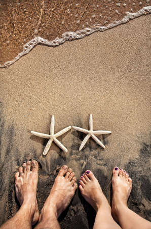 Legs of young couple standing on the beach near two starfishes on the sand with ocean near by