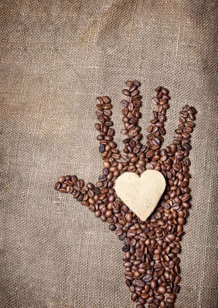 Hand from coffee beans holding heart shape cookie on textured brown sackの写真素材