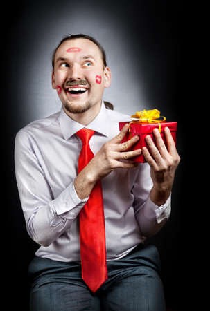 Happy Man with moustache and marks from red lipstick on his face holding red box present at black backgroundの写真素材