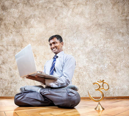 Happy Indian businessman doing yoga and typing on his laptop with om statue near by in the office at brown backgroundの写真素材