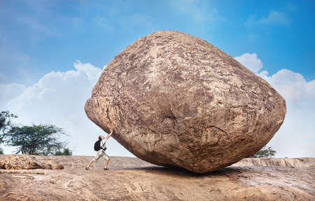 Man with backpack pushing a huge stone in Mamallapuram cave complex, Tamil Nadu, India