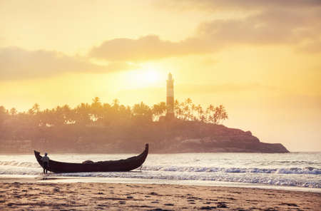 Indian fisherman in silhouette with fishing boat on the beach in the morning at lighthouse background in Kovalam, Kerala, India  の写真素材