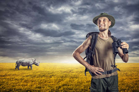 Man with binocular and rhino behind in grassland at sunset overcast skyの写真素材