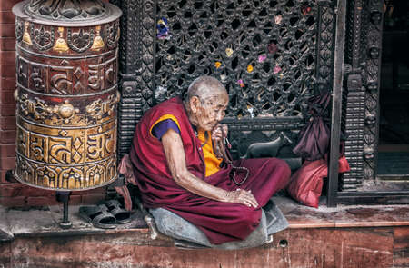 BODNATH, KATHMANDU, NEPAL - APRIL 7, 2014: Old woman in red Buddhist robe sitting near small temple with prayer wheel at Bodnath stupaのeditorial素材
