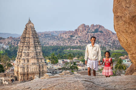 HAMPI, KARNATAKA, INDIA - MARCH 11, 2013: Indian man with his daughter in red dress walking on Hemakuta hill at Virupaksha templeのeditorial素材