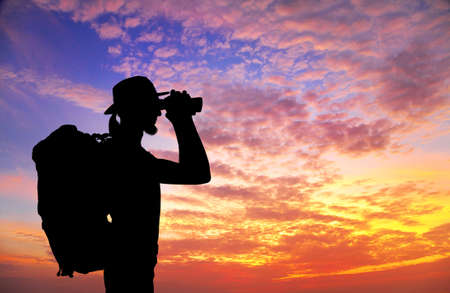 Man with backpack looking through the binocular in silhouette at sunset sky backgroundの写真素材