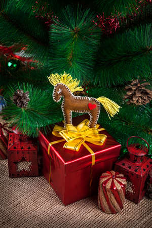 Horse toy from felt on the red box with yellow bow and lanterns, candle near Christmas treeの写真素材