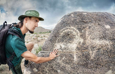 Man in hat with backpack touching the stone with ancient petroglyph of goat の写真素材