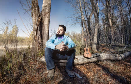 Man in blue shirt with bow tie and ukulele on the tree trunk in the forest