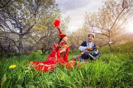 Kazakh man playing dombra and singing the song for woman in red on the green grass in Almaty, Kazakhstan, Central Asiaの写真素材