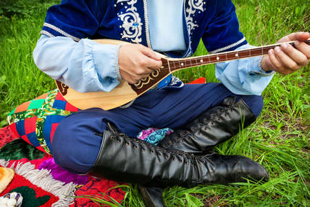 Man in traditional Kazakh costume playing dombra instrument on the green grass in apple garden of Almaty, Kazakhstan, Central Asiaの写真素材