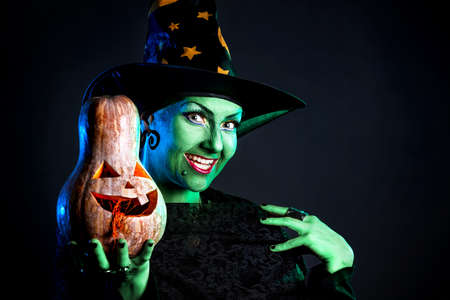 Witch with green skin holding carved Halloween pumpkin at dark backgroundの写真素材