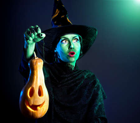 Witch with green skin holding carved Halloween pumpkin lantern at dark backgroundの写真素材