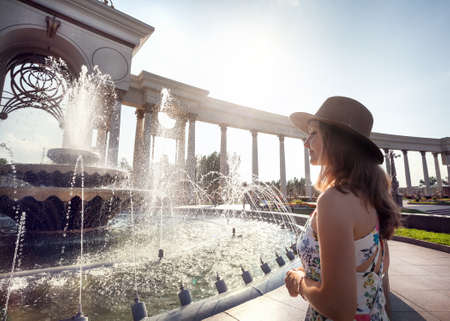 Woman in white dress and hat near the fountain in the park with big arch in Almaty, Kazakhstanの写真素材