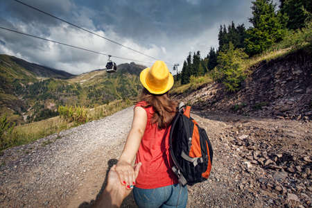 Tourist woman with backpack and yellow hat holding man by hand and walking down the road near cableway in the mountain valleyの写真素材