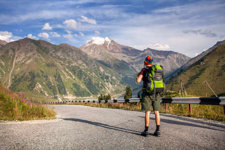 Tourist with backpack and rainbow hat walking down the road in the mountains near Big Almaty Lake in...