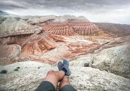 Tourist legs with gray shoes on Desert Mountains with red and white layers at cloudy sky background
