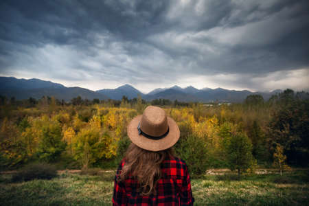 Woman in red checked shirt and hat looking at autumn forest and mountains background. Travel concept.の写真素材