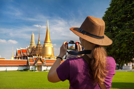 Woman tourist in hat taking a picture of Temple of the Emerald Buddha with Golden Stupa in Bangkok の写真素材
