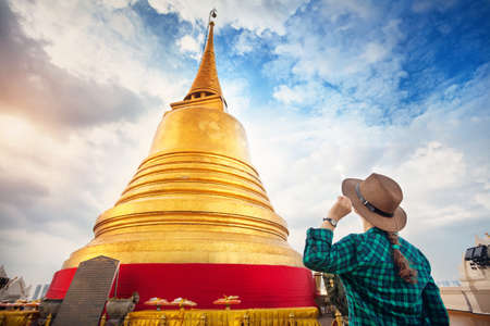 Tourist woman in hat and green checked shirt looking at big golden Stupa in Wat Saket temple in Bangkok, Thailandの写真素材