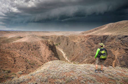 Man with travel backpack in hat and green shirt on top of the canyon looking to the view of valley and storm dark skyの写真素材