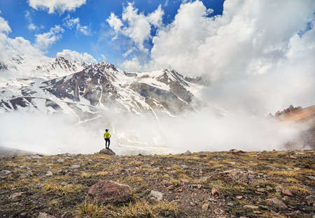 Hiker in yellow shirt standing on the stone with snowy mountains at overcast sky background
