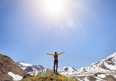 Hiker in yellow shirt with backpack standing on the rock with rising hands enjoying the view of snowy mountains at sunny day skyの写真素材