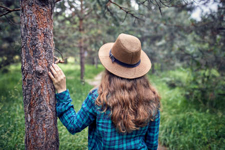 Young woman in brown hat and green checked shirt with long hair touching the pine tree in the forest