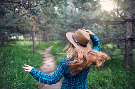 Young woman in brown hat and green checked shirt with long hair running under the in the pine tree forestのeditorial素材