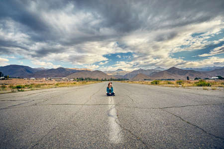 Woman in hat and checked shirt sitting with cross leg on the wide asphalt road with mountains and cloudy sky backgroundの写真素材