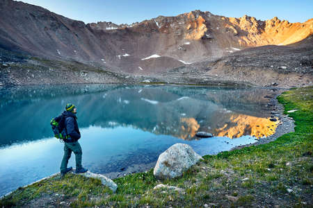 Bearded man in green hat and backpack looking at mountain lake at sunriseの写真素材