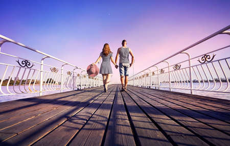 Woman in striped dress with red hat and man in stripped shirt going on the pier near lake at the pink sunsetの写真素材