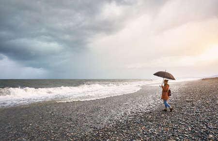 Woman with umbrella and backpack walking at stormy sea shore with overcast sky in Batumi, Georgiaの写真素材