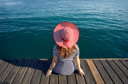 Woman in hat and striped dress sitting on pier and looking the emerald blue sea. Summer and Sea concept.の写真素材